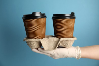 Woman in latex gloves holding cardboard holder with takeaway paper coffee cups against light blue background, closeup