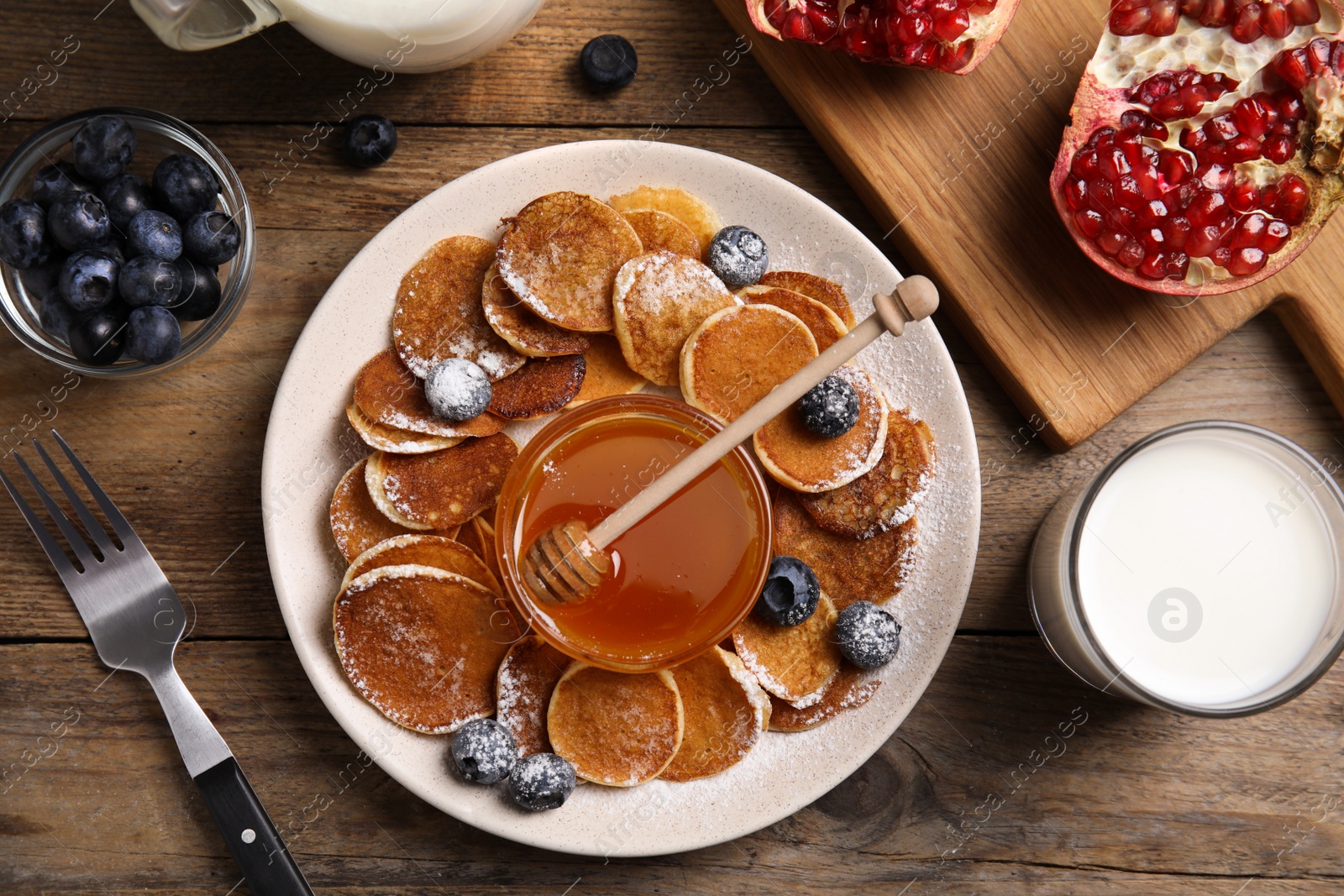 Photo of Delicious mini pancakes cereal with blueberries and honey served on wooden table, flat lay
