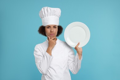 Photo of Thoughtful female chef in uniform holding plate on light blue background