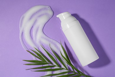 Photo of Bottle with cleansing foam and green leaves on violet background, flat lay. Cosmetic product