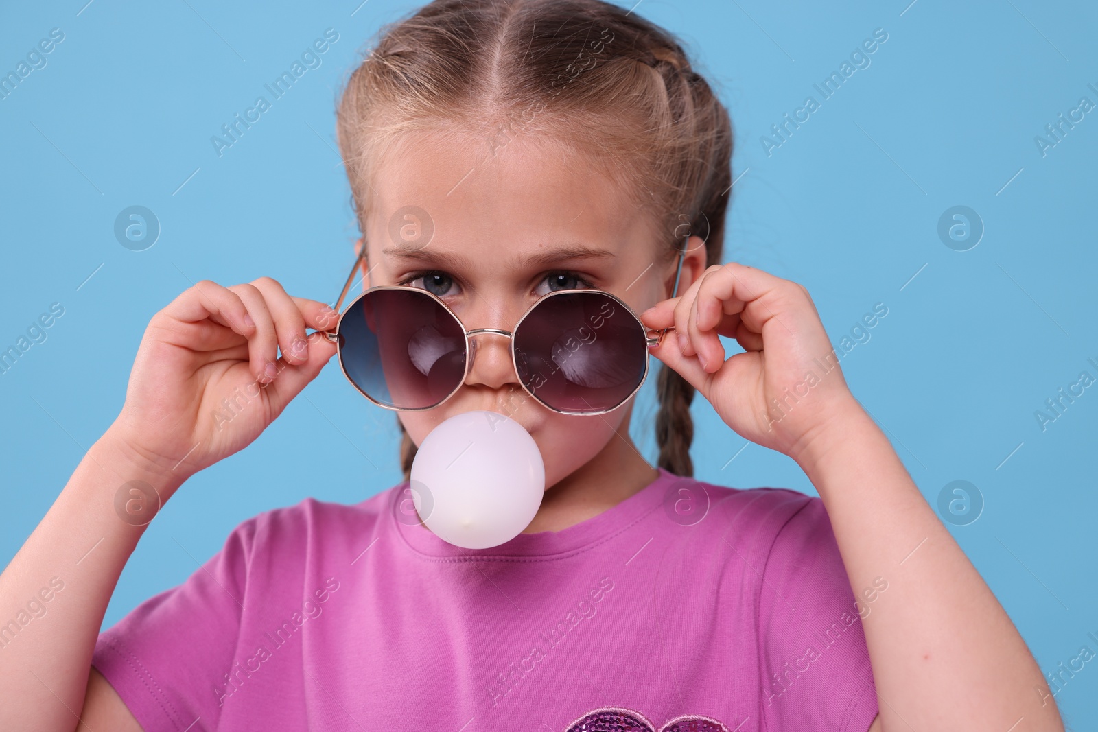 Photo of Girl in sunglasses blowing bubble gum on light blue background