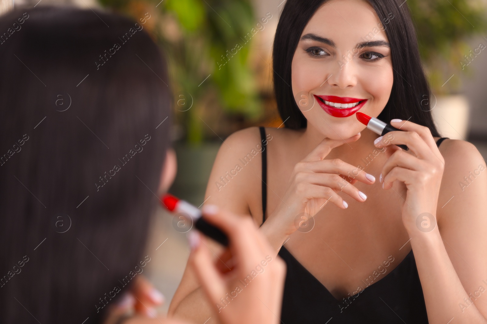 Photo of Young woman applying beautiful red lipstick in front of mirror indoors