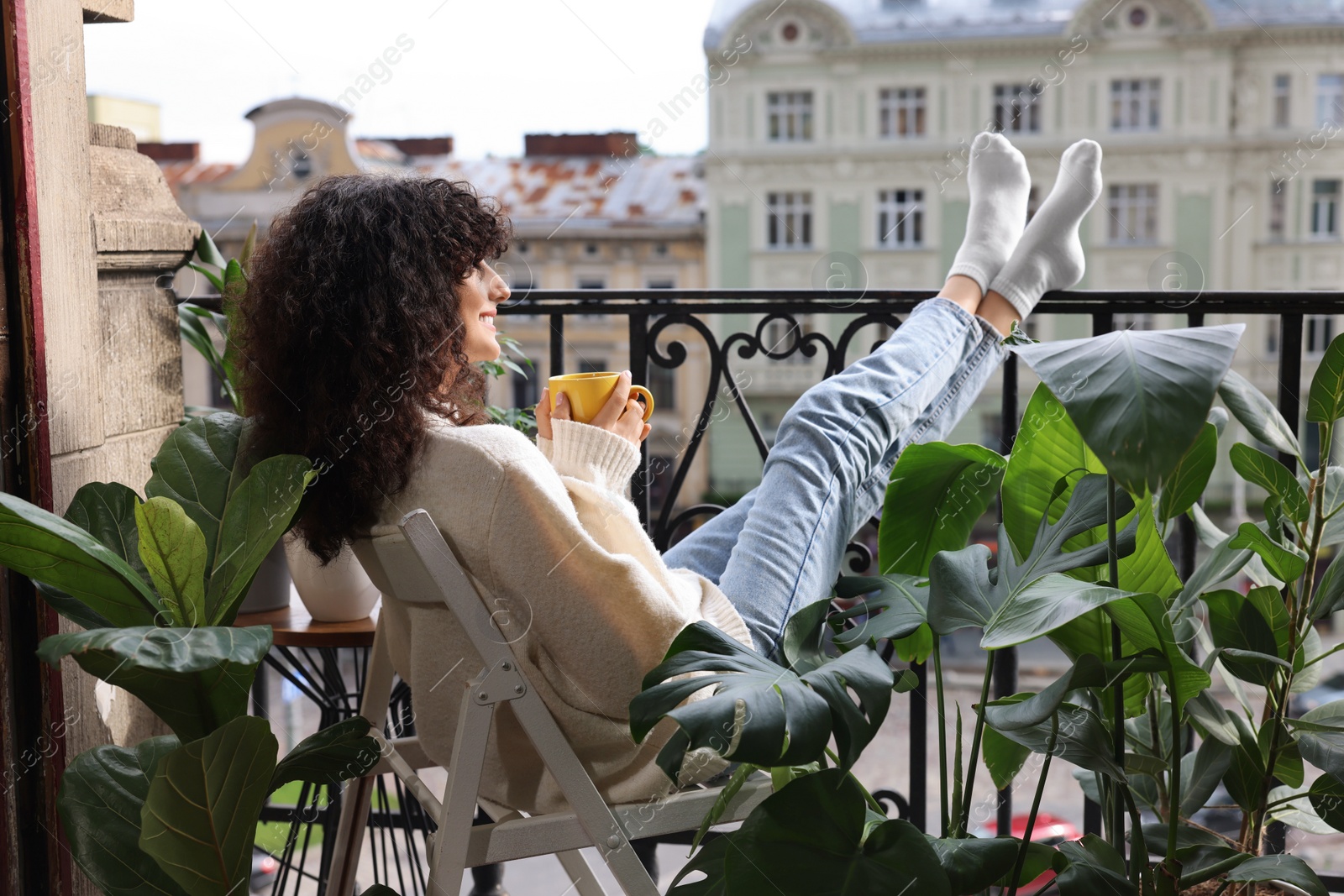 Photo of Young woman with cup of tea relaxing in chair surrounded by green houseplants on balcony