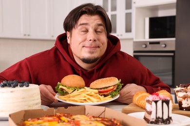 Photo of Happy overweight man holding plate with tasty burgers and French fries at table in kitchen