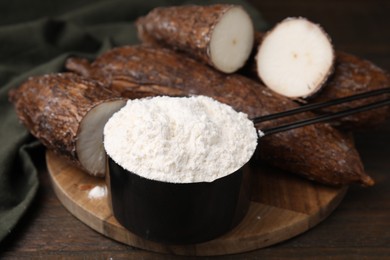Scoop with cassava flour and roots on wooden table, closeup