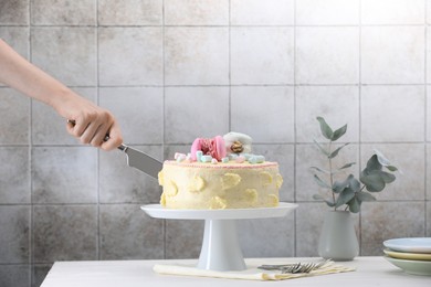 Woman cutting delicious cake decorated with macarons and marshmallows at white wooden table, closeup. Space for text