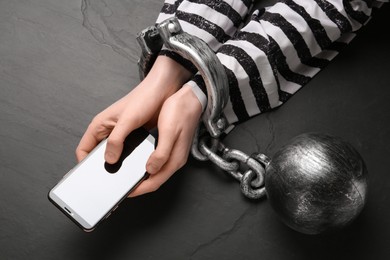 Photo of Prisoner shackled with ball and chain holding smartphone at black table, top view. Internet addiction