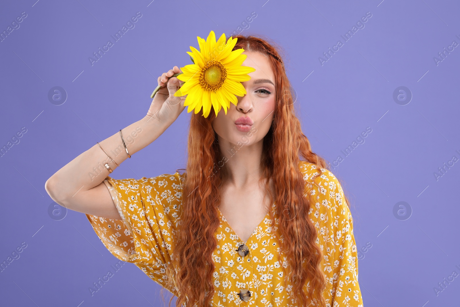 Photo of Beautiful young hippie woman covering eye with sunflower and sending air kiss on violet background