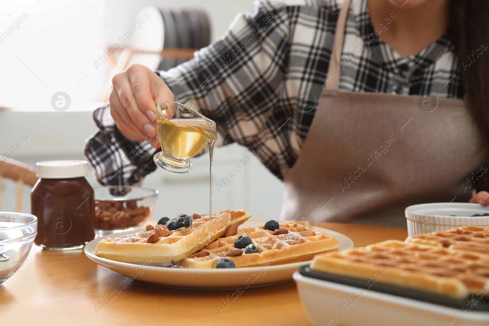 Photo of Woman pouring honey onto delicious Belgian waffles  at wooden table in kitchen, closeup