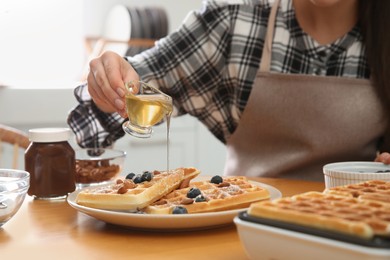 Woman pouring honey onto delicious Belgian waffles  at wooden table in kitchen, closeup