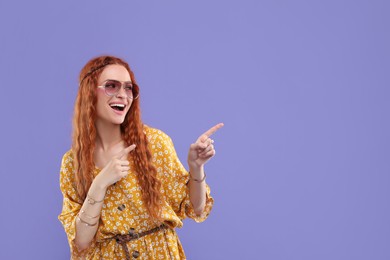 Stylish young hippie woman in sunglasses pointing at something on violet background, space for text