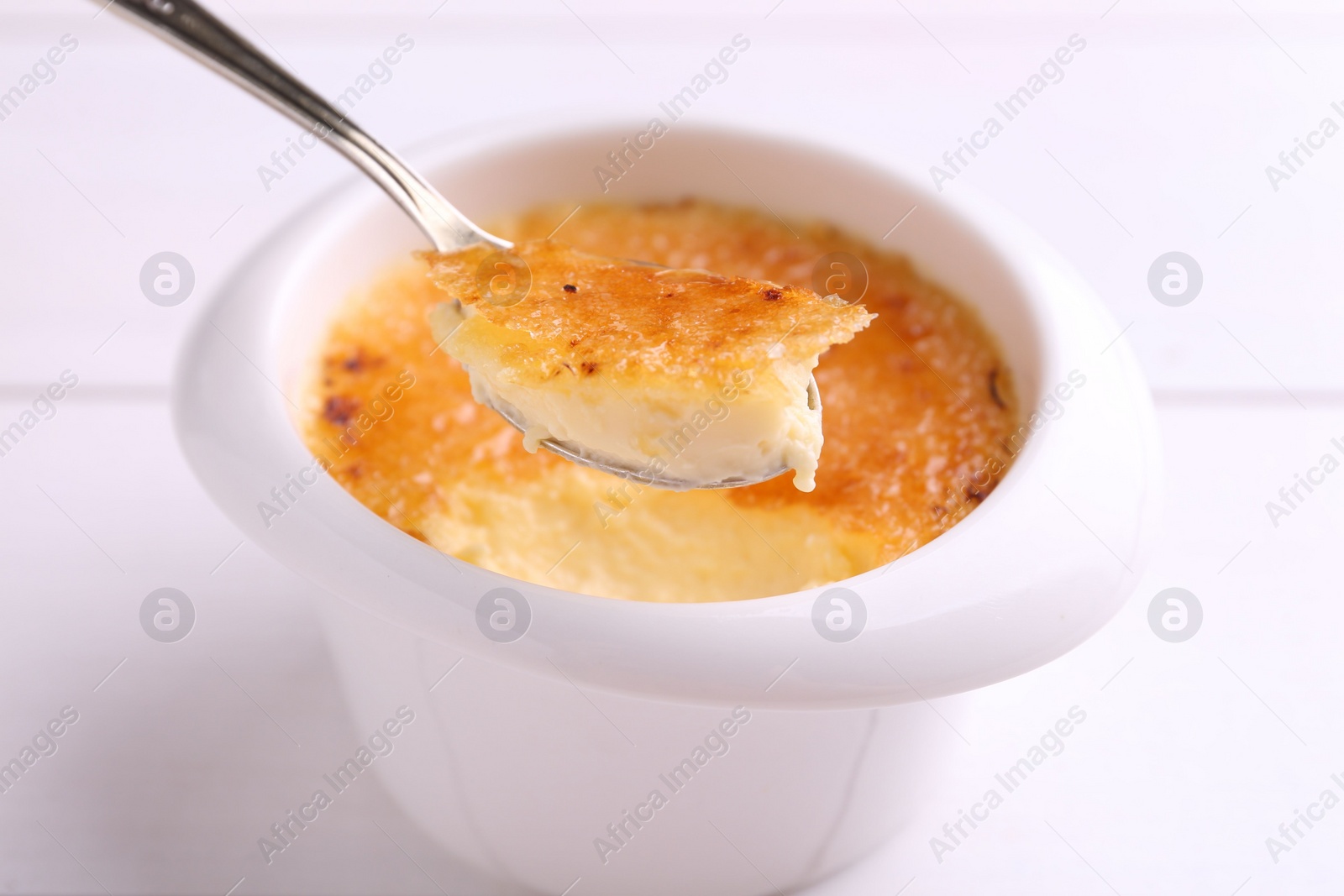 Photo of Taking delicious creme brulee with spoon from bowl at white table, closeup