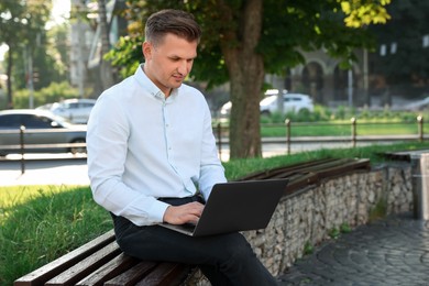 Handsome man using laptop on bench outdoors. Space for text