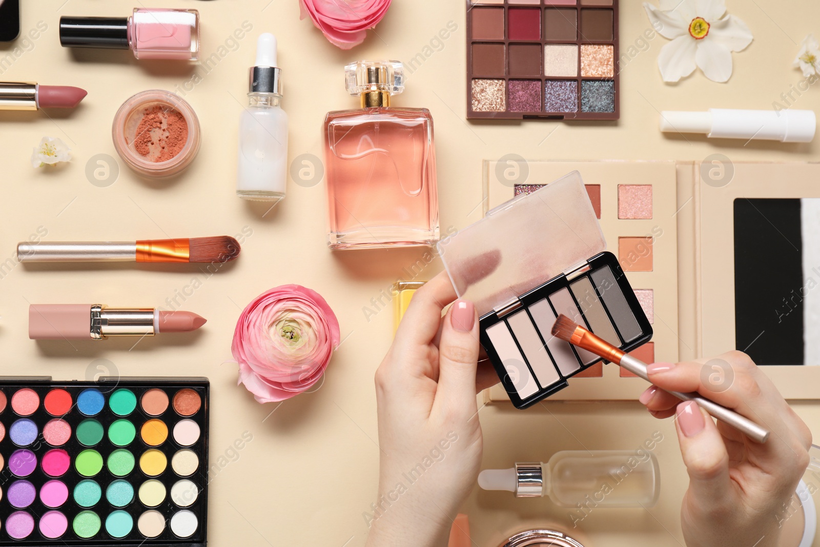 Photo of Woman applying eye shadow palette on brush, closeup. Different makeup products and beautiful spring flowers on beige background, top view