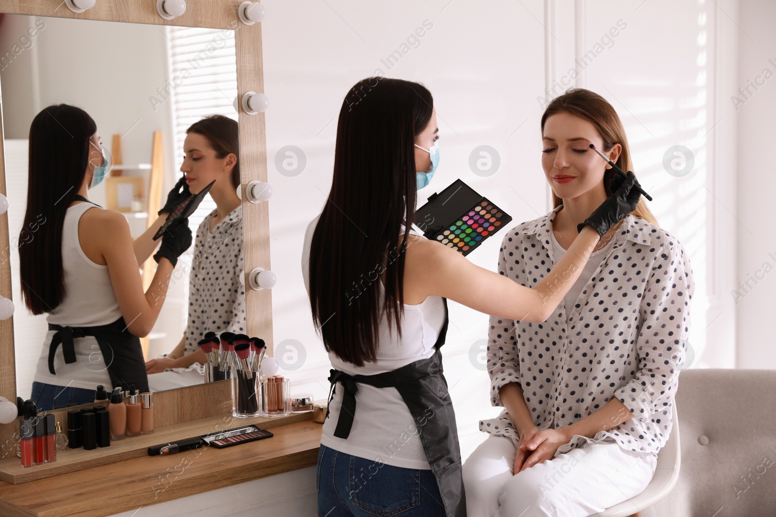 Photo of Makeup artist in protective mask and gloves working with woman indoors. Preventive measures during COVID-19 pandemic