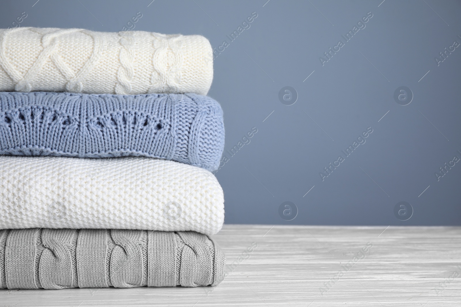 Photo of Stack of folded knitted sweaters on white wooden table against grey background, closeup. Space for text