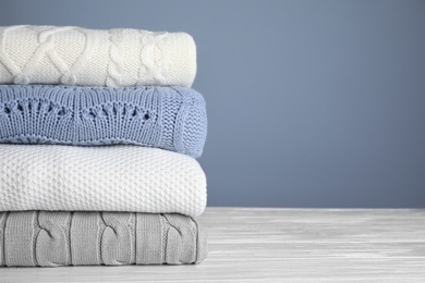 Stack of folded knitted sweaters on white wooden table against grey background, closeup. Space for text