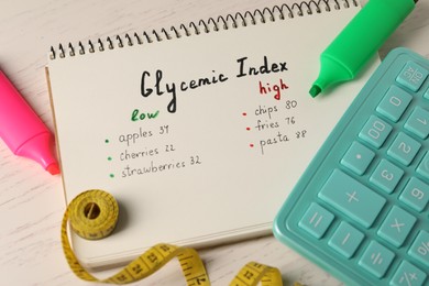 List with products of low and high glycemic index in notebook, markers, measuring tape and calculator on white wooden table, above view