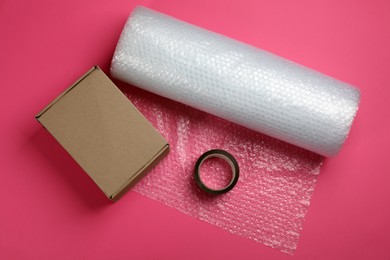 Photo of Bubble wrap roll, cardboard box and tape on pink background, flat lay