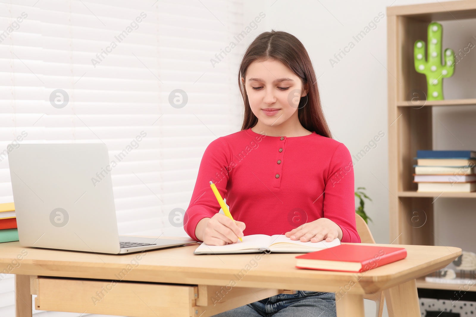 Photo of Cute girl writing in notepad near laptop at desk in room. Home workplace