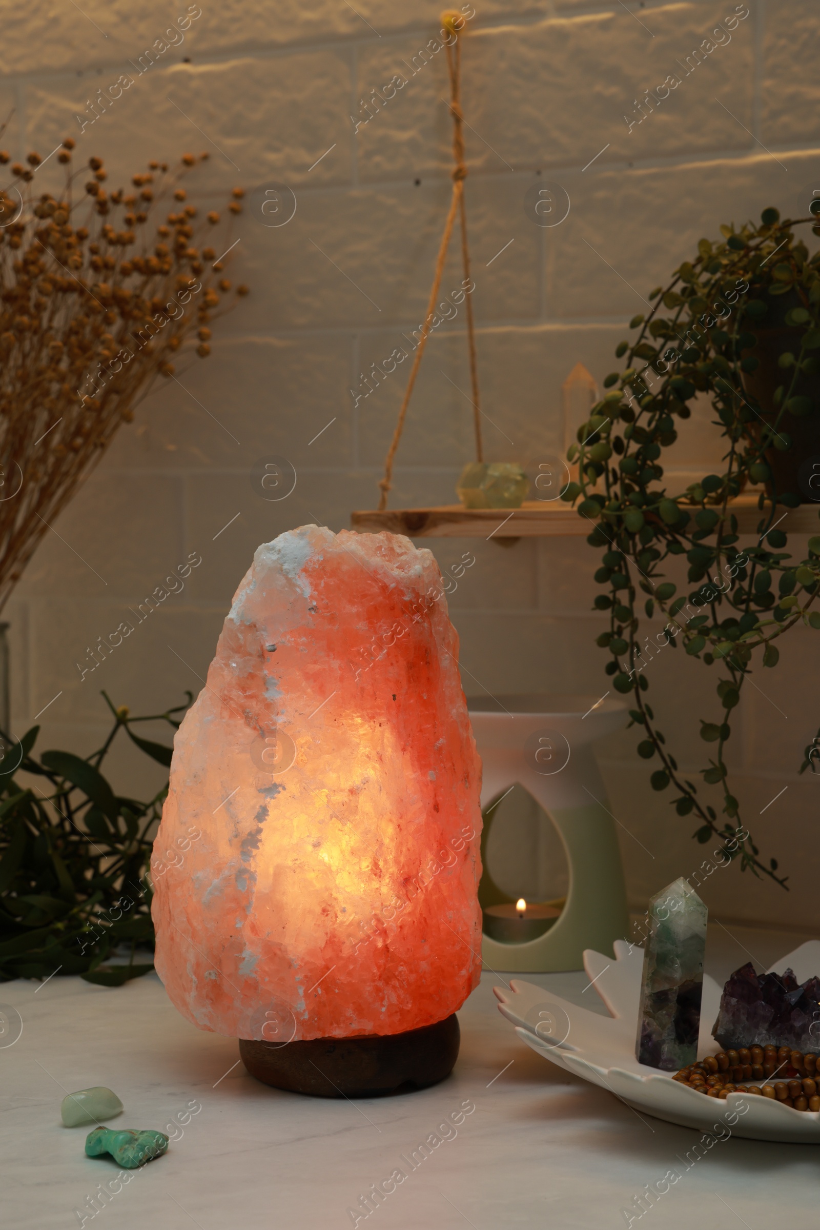 Photo of Himalayan salt lamp, crystals and oil diffuser on white table near brick wall
