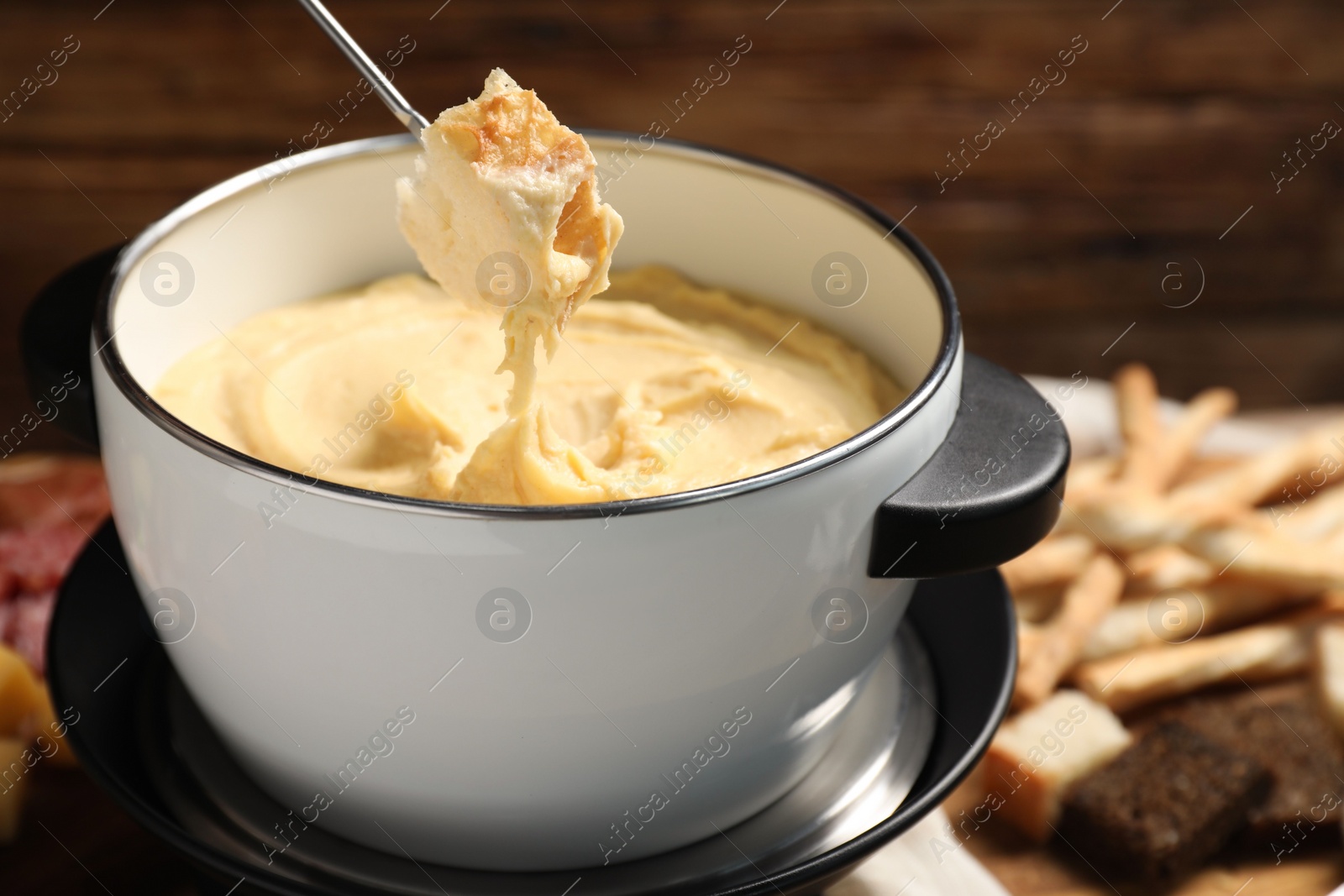 Photo of Dipping piece of bread into fondue pot with melted cheese on blurred background, closeup