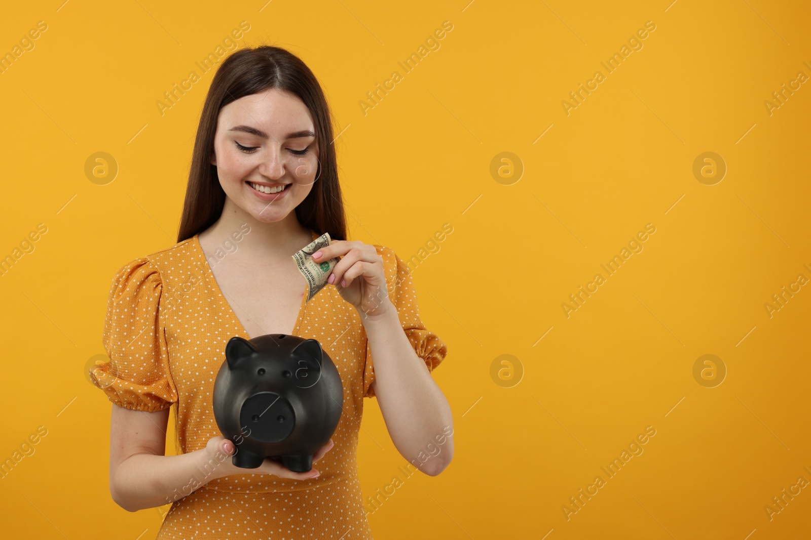 Photo of Happy woman putting dollar banknote into piggy bank on orange background, space for text