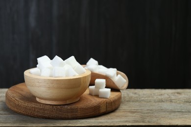 Photo of White sugar cubes in bowl and scoop on wooden table against dark background, closeup. Space for text