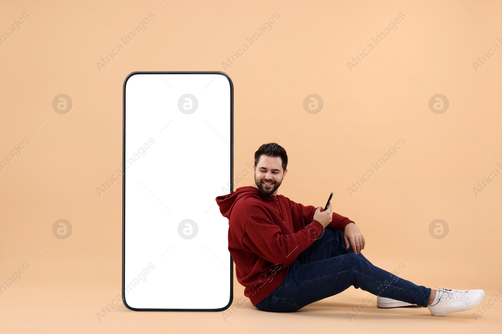 Image of Man with mobile phone sitting near huge device with empty screen on dark beige background. Mockup for design