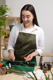 Photo of Young woman planting vegetable seeds into plastic pots with soil at wooden table indoors