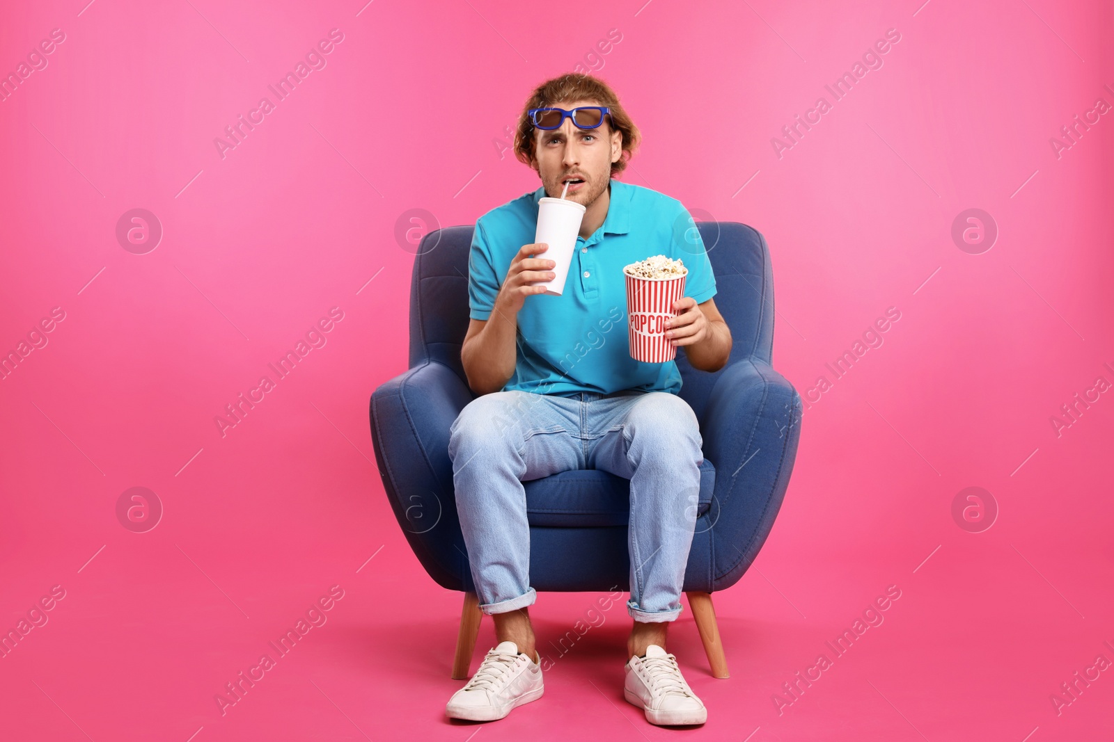 Photo of Emotional man with 3D glasses, popcorn and beverage sitting in armchair during cinema show on color background