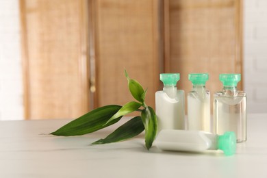 Photo of Mini bottles of cosmetic products and green branch on white table against blurred background. Space for text