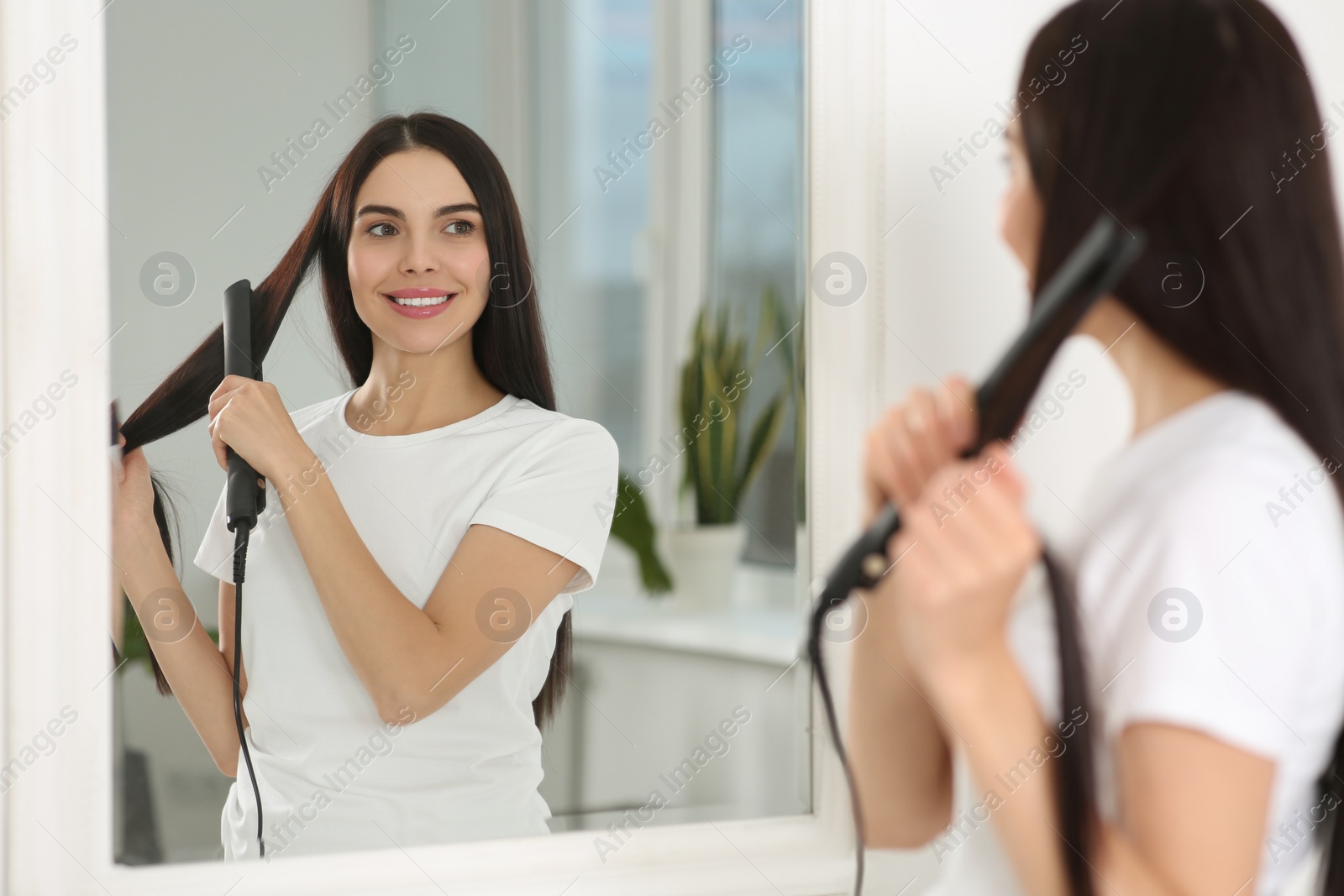 Photo of Beautiful happy woman using hair iron near mirror in room