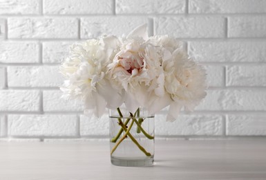 Photo of Beautiful peonies in glass vase on white table near brick wall