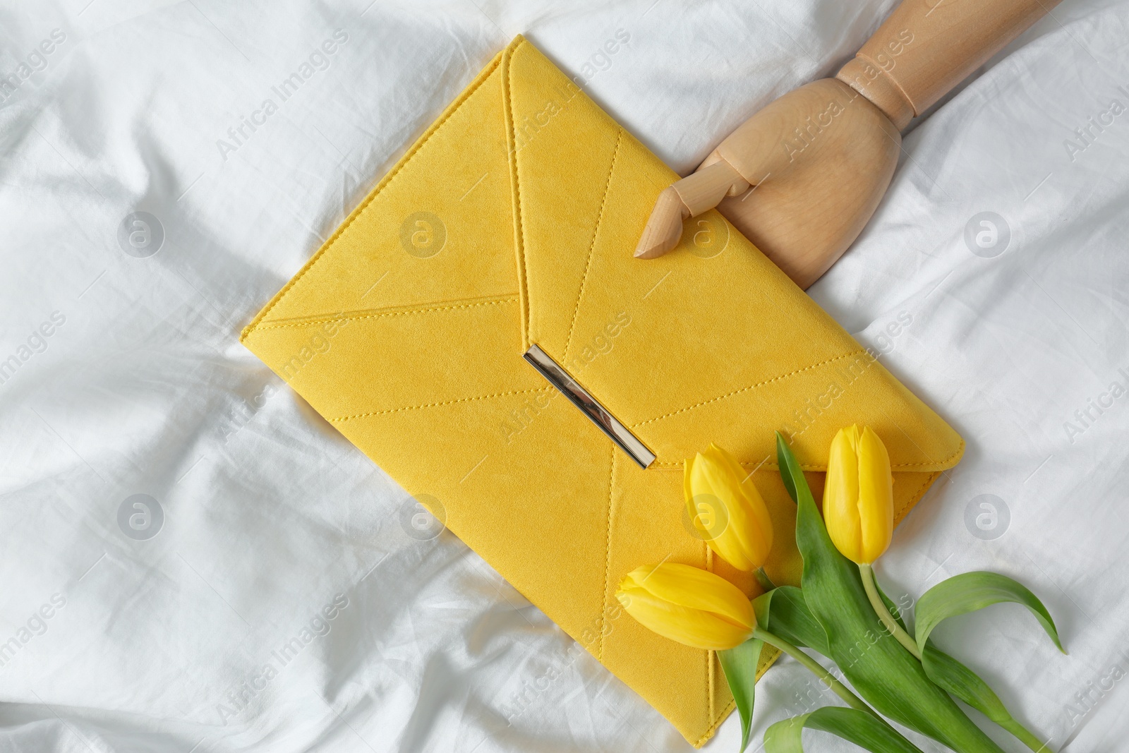 Photo of Stylish envelope bag with spring tulips and wooden mannequin hand on crumpled sheet, flat lay