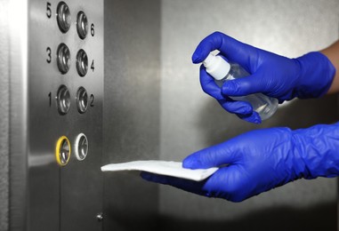 Woman with detergent spray and paper napkin cleaning elevator`s panel, closeup