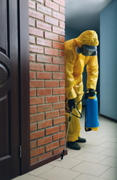 Photo of Pest control worker in protective suit spraying pesticide indoors