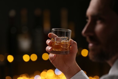 Photo of Man holding glass of whiskey with ice cubes against blurred lights, selective focus