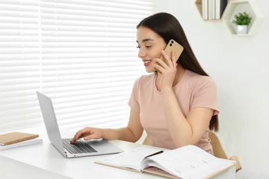 Home workplace. Happy woman talking on smartphone near laptop at white desk in room