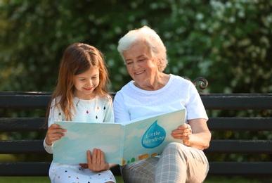 Photo of Little girl and her grandmother reading book on bench in park