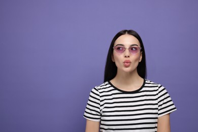 Photo of Beautiful young woman in stylish sunglasses blowing kiss on purple background. Space for text