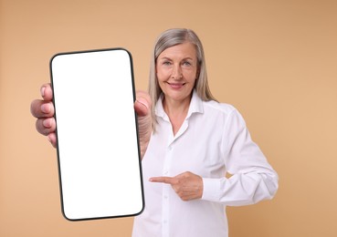 Happy mature woman pointing at mobile phone with blank screen on beige background. Mockup for design