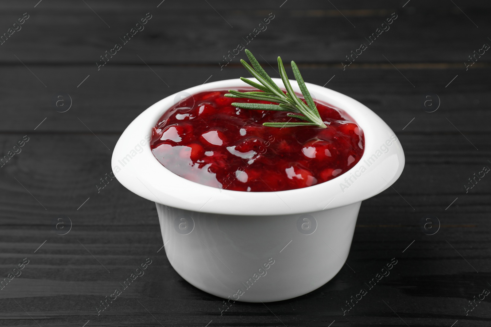 Photo of Fresh cranberry sauce in bowl and rosemary on black wooden table, closeup