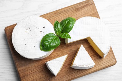 Photo of Tasty cut and whole brie cheeses with basil on white wooden table, top view