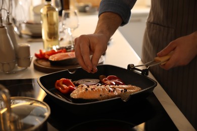 Man pouring pepper into frying pan with tasty salmon steak and vegetables, closeup