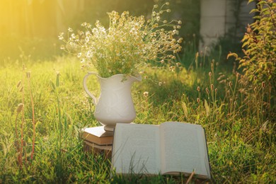 Photo of Open book with ceramic jug with chamomiles on green grass outdoors