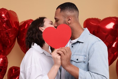 Photo of Lovely couple kissing behind red paper heart on beige background. Valentine's day celebration