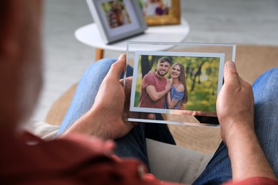 Photo of Senior man holding frame with photo of young couple at home, closeup