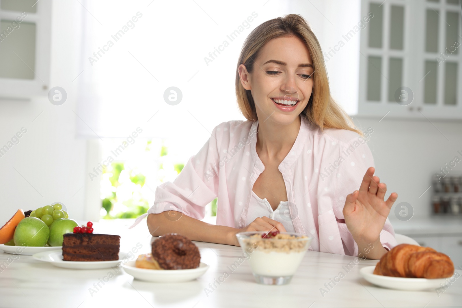 Photo of Woman choosing between sweets and healthy food at white table in kitchen