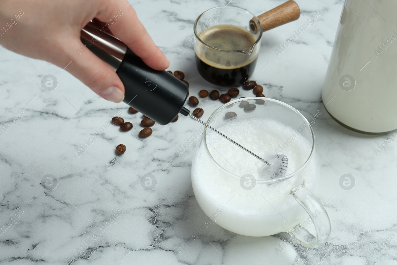 Photo of Woman whisking milk in cup with mini mixer (milk frother) at white marble table, closeup